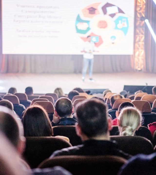 Audience attentively watching a presentation in a conference setting, with stage lights and speaker visible.